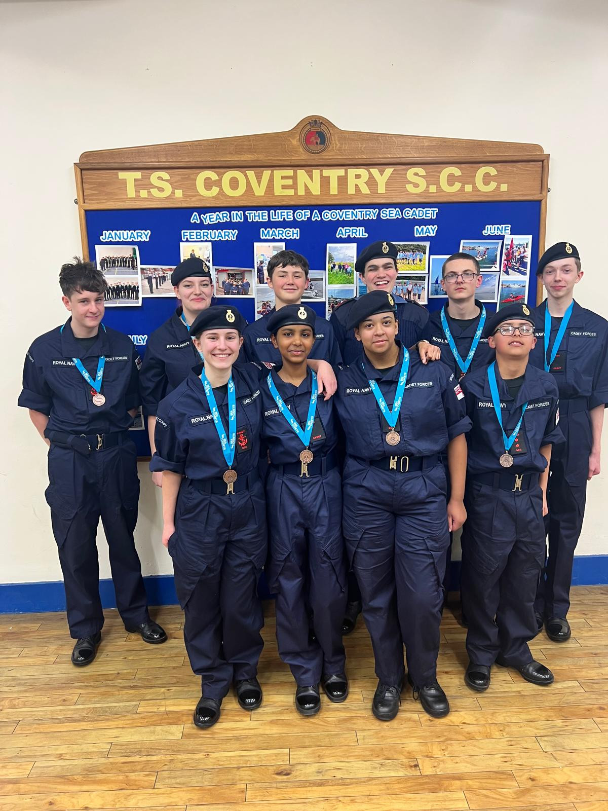 The Coventry Drill team in their PCS with their bronze medals stood as a group in front of a display board